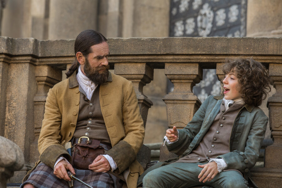 Murtagh and Fergus sitting outside an ornate stone building in Paris. Fergus is laughing and talking animatedly, while Murtagh looks on with a furrowed brow.