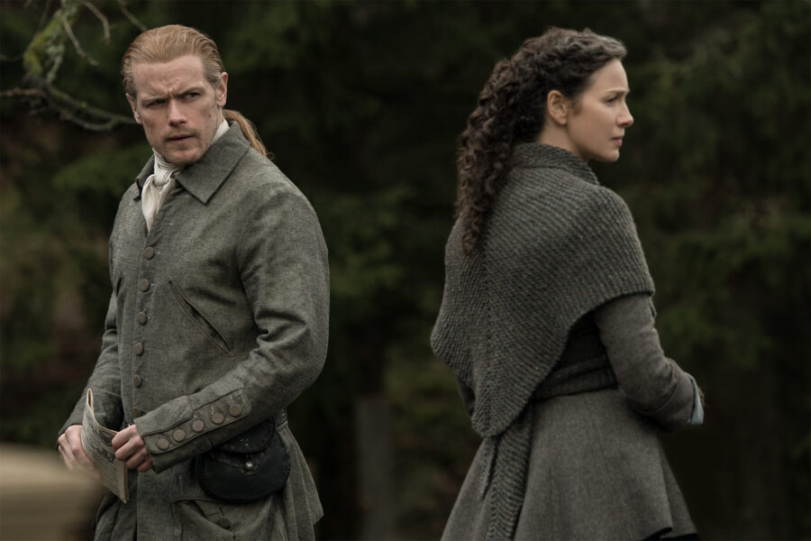 Jamie and Claire stand back to back pensively. Jamie is holding the newspaper.