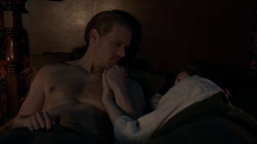 Jamie and Claire laying in bed holding hands.