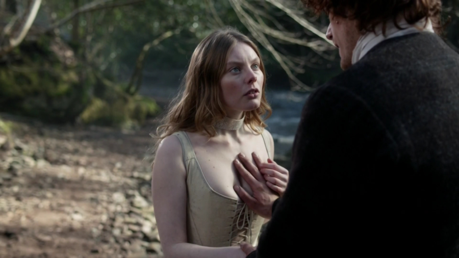 Screencap from season one episode The Reckoning. Leghaire has placed Jamie’s hand on her exposed chest as they stand on the bank of a creek.